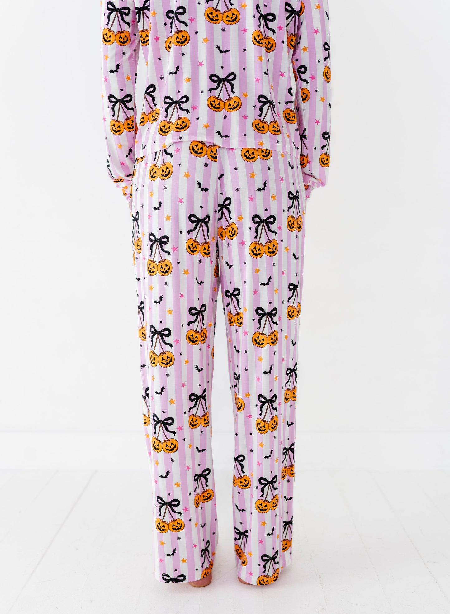 A person wearing a matching long-sleeve pajama set with a Halloween print, shown from the back against a plain white studio background. The pajamas are light pink with an allover pattern of orange jack-o'-lanterns, black bows, and small black bats.