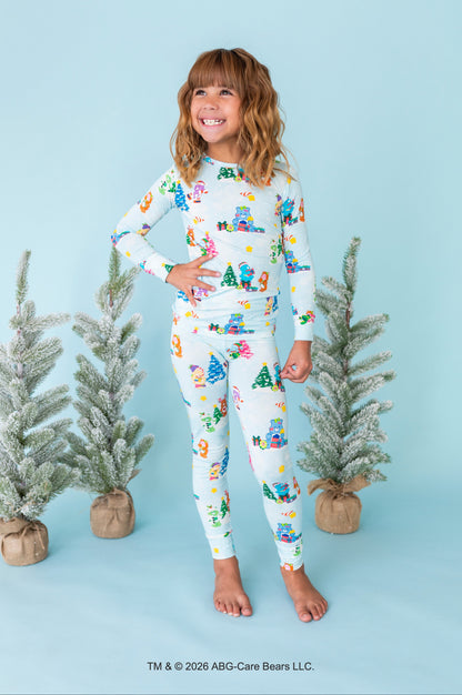 A young girl stands barefoot against a light blue studio backdrop, wearing a matching long-sleeve pajama top and leggings set with a colorful Christmas tree and snowman print on a white base. Small potted frosted trees are placed around her, and a copyright notice for ABC-Care Bears LLC appears at the bottom.