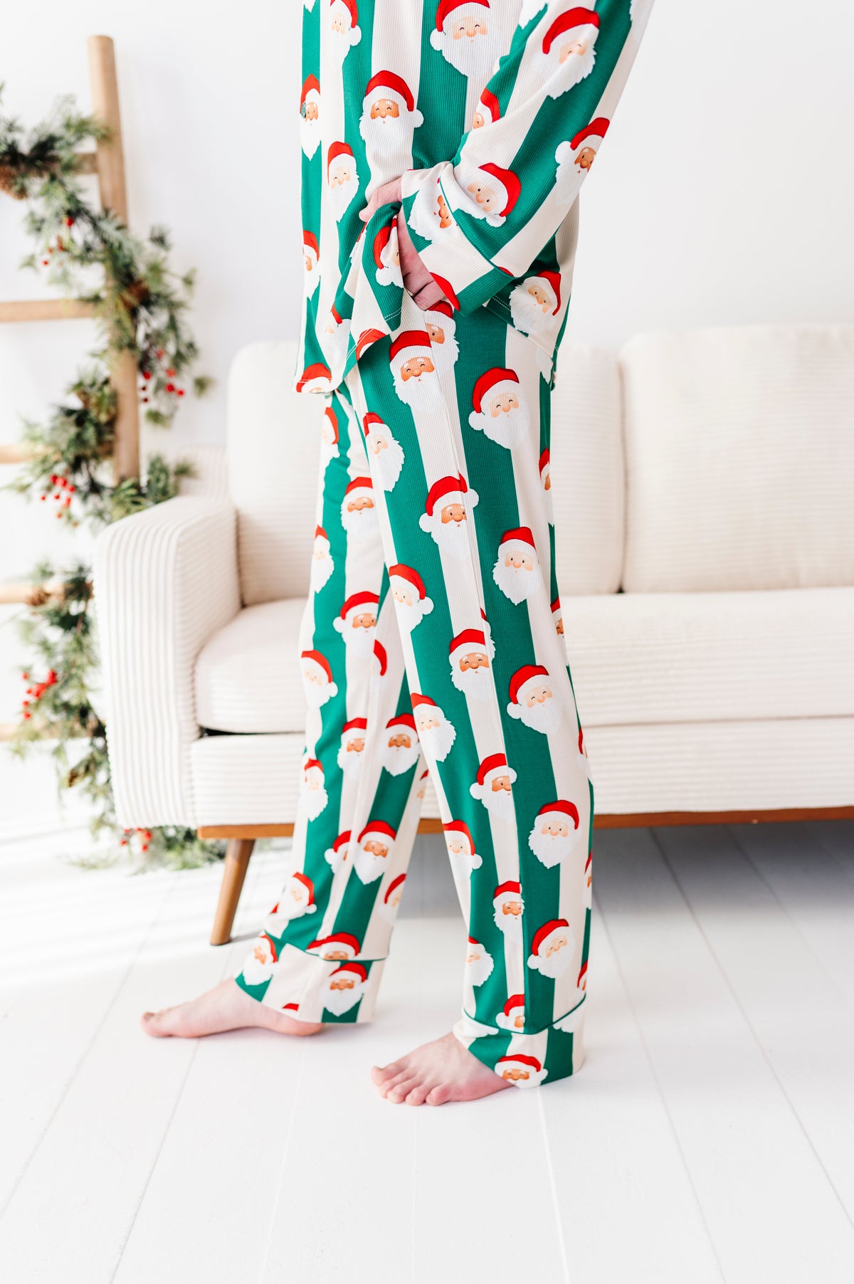 A person wearing green and white Santa Claus patterned bamboo pajamas, standing barefoot in a bright living room with a white sofa and holiday decor.