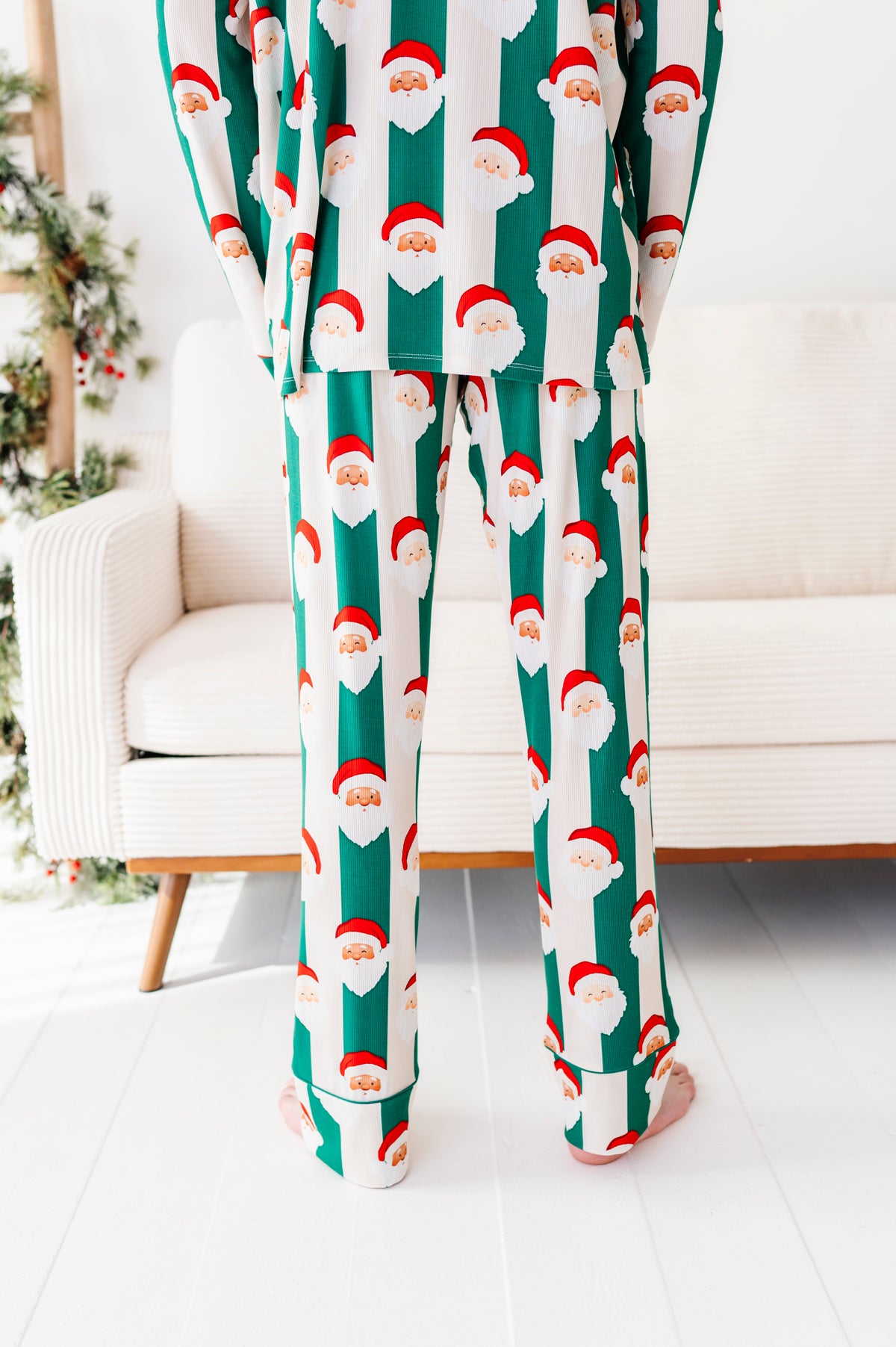 A person wearing a matching Christmas bamboo pajama set with a green and white striped pattern and Santa Claus faces, standing barefoot in a bright living room.