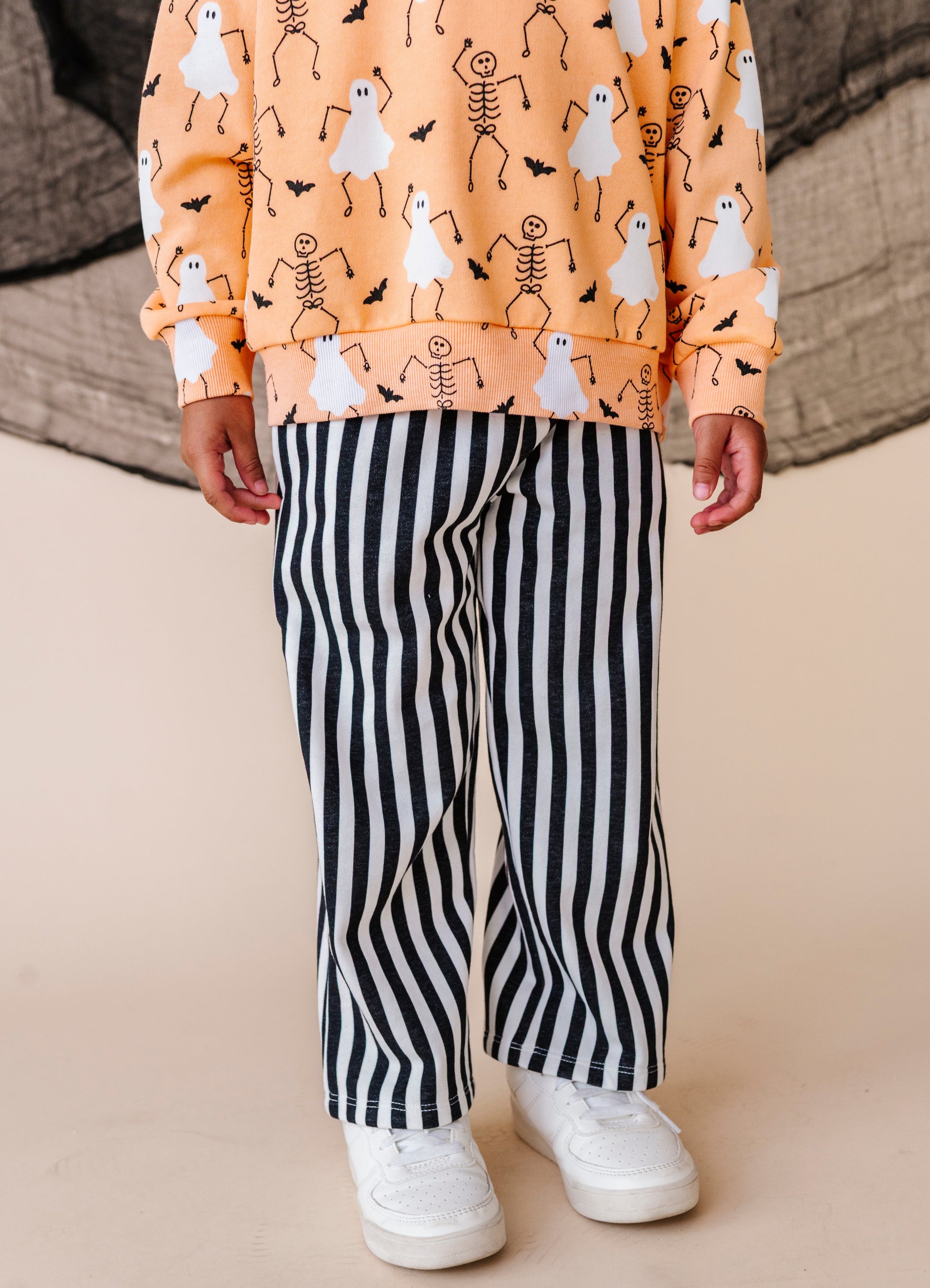 Child wearing a Halloween-themed sweatshirt with ghosts and skeletons and black and white striped pants on a beige floor.