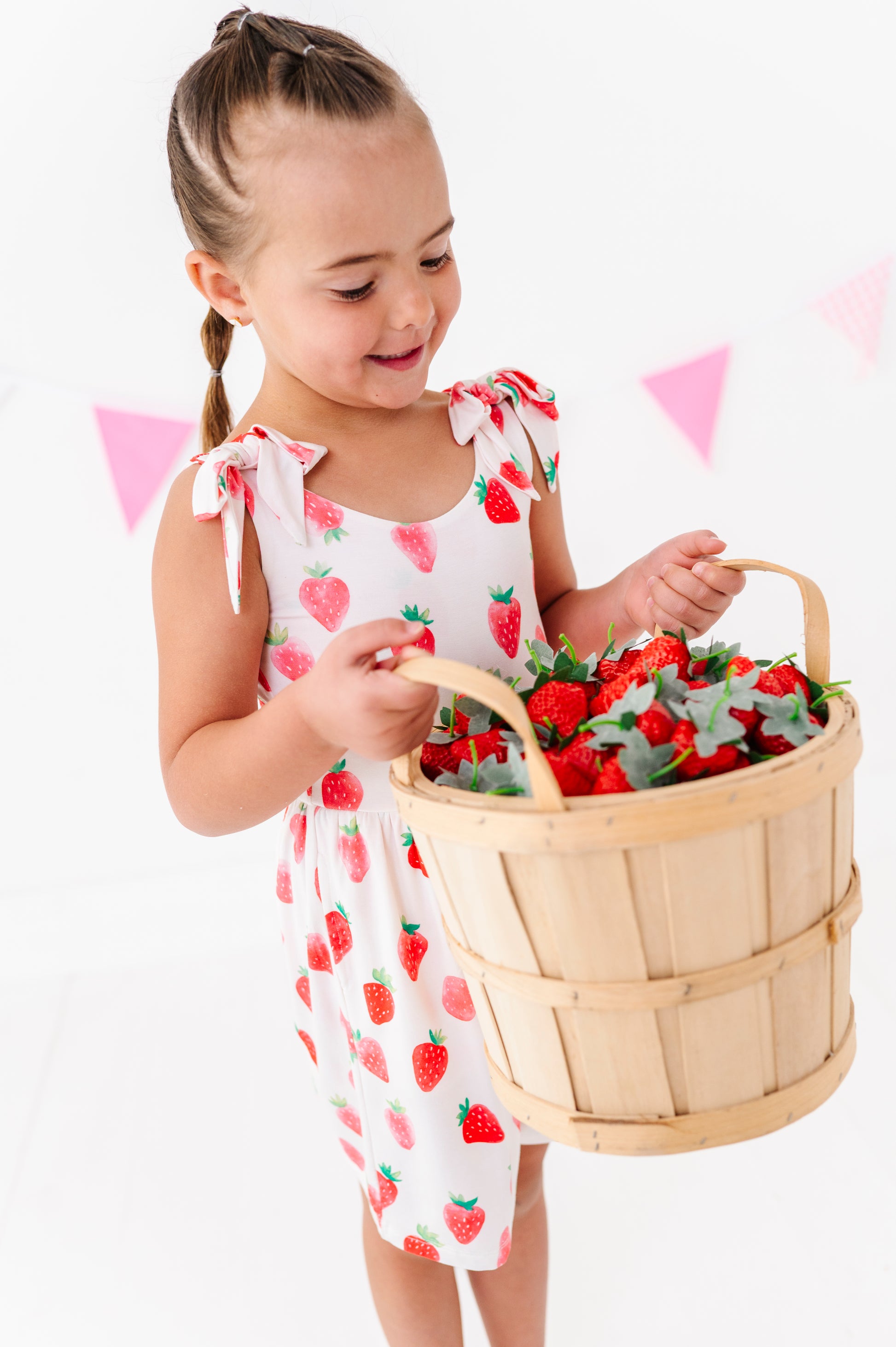Young girl in a strawberry outfit holding a basket of strawberries against a white background with pink triangle flags.