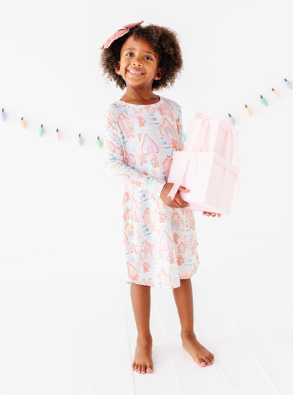 A little girl wearing a Christmas gingerbread-themed bamboo nightgown, standing in a bright white room and holding a stack of pink wrapped presents