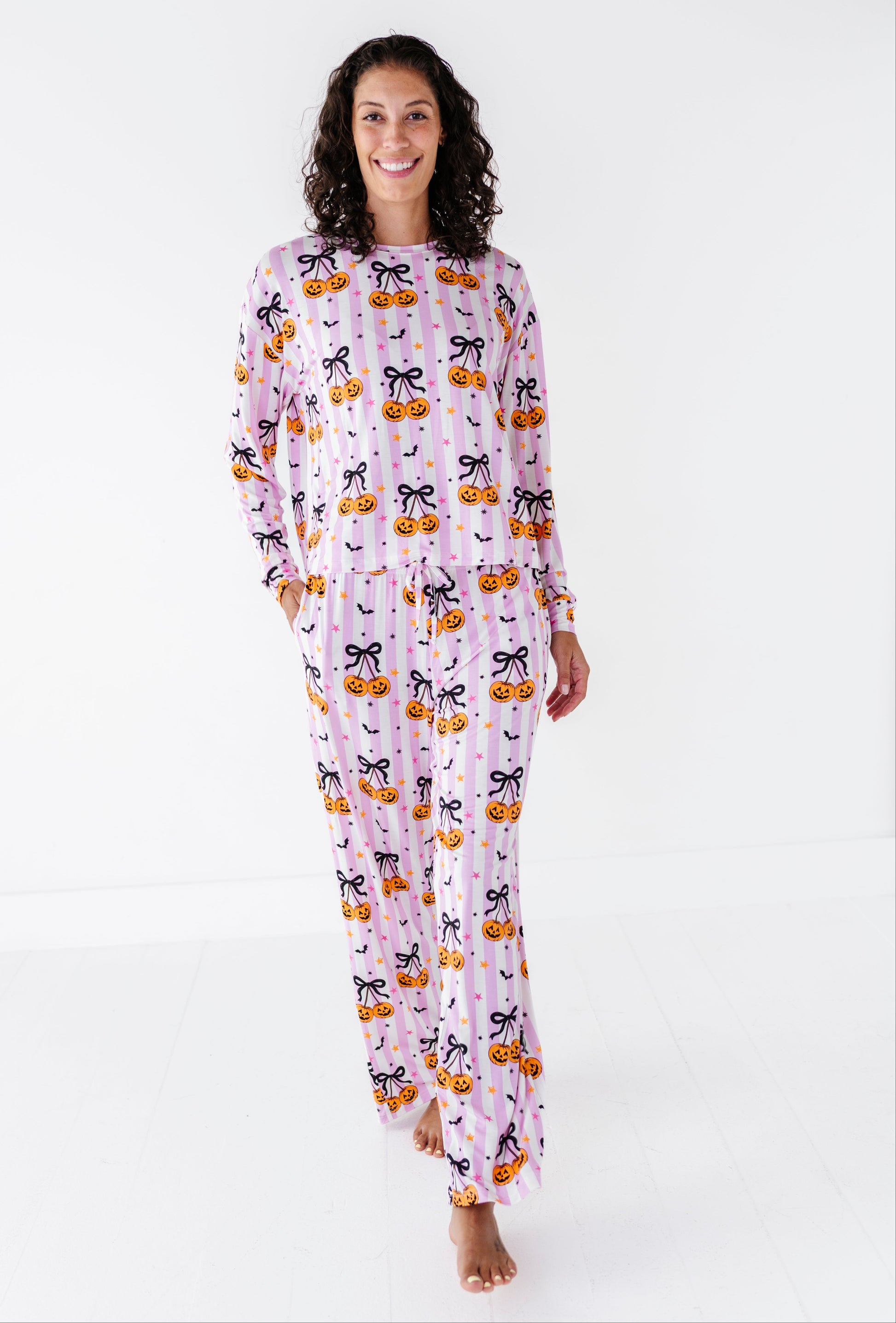 A woman modeling a long-sleeve pajama set with a Halloween pumpkin and cat print, standing against a plain white studio background.