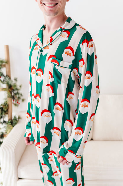 A man wearing a long-sleeve bamboo pajama shirt and matching pants with a green and white striped pattern featuring Santa Claus faces, standing in a bright living room.