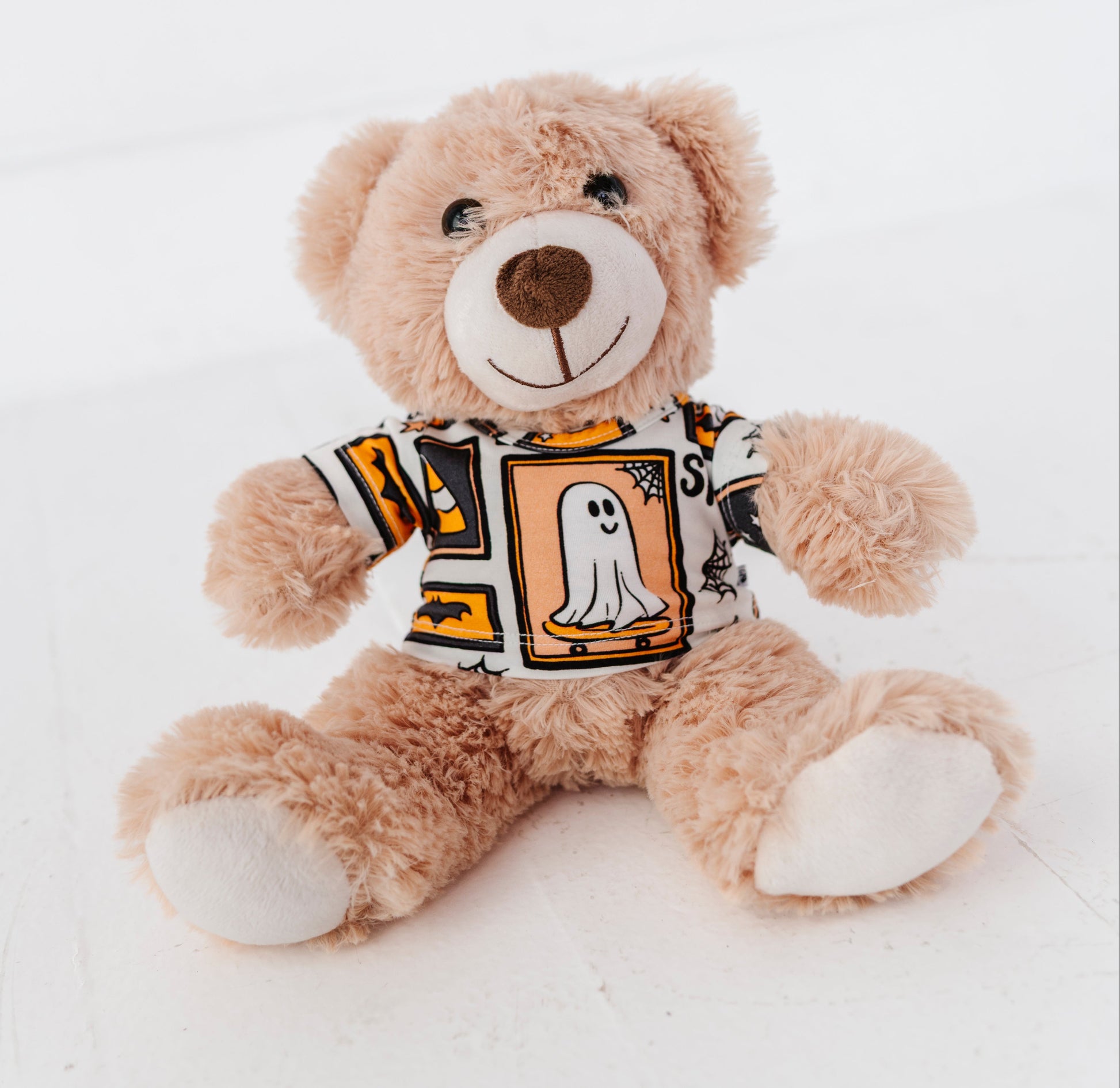 A plush teddy bear sitting on a white surface, wearing a short-sleeve Halloween-themed shirt with a ghost graphic and orange, black, and white patterned design.