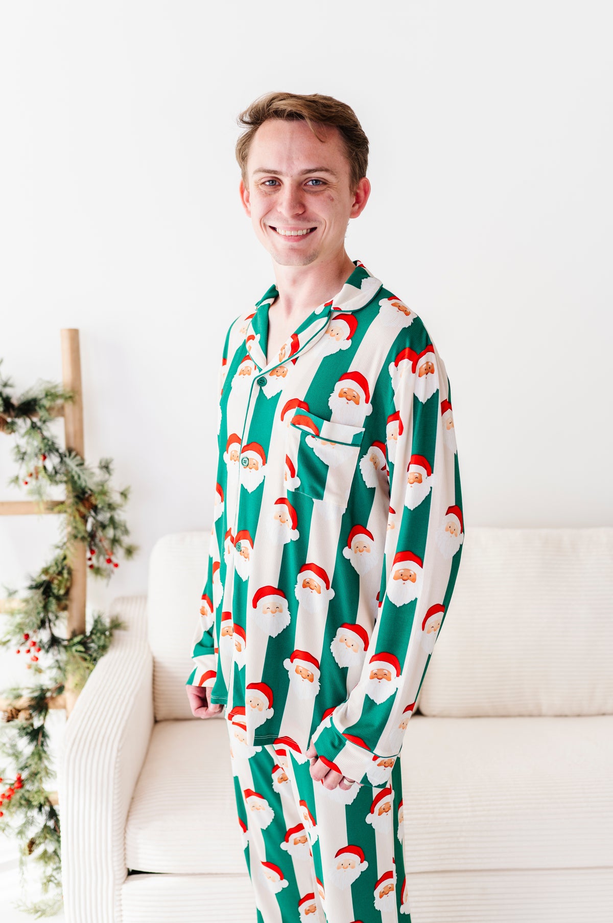 A man wearing a green and white long-sleeve bamboo pajama set with a repeating Santa Claus face pattern, standing in front of a white couch with holiday greenery on the left.