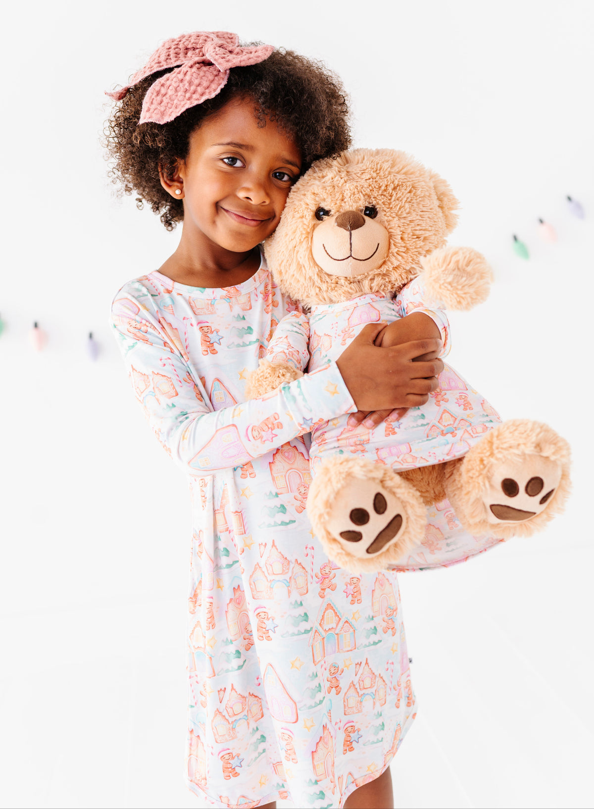 A little girl wearing a Christmas gingerbread-themed bamboo nightgown, standing in a bright white room and holding a stuffed teddy bear wearing a matching bamboo nightgown 