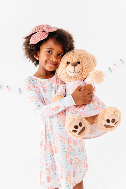 A little girl holding a teddy bear. They are wearing matching Christmas gingerbread village-themed bamboo gowns 