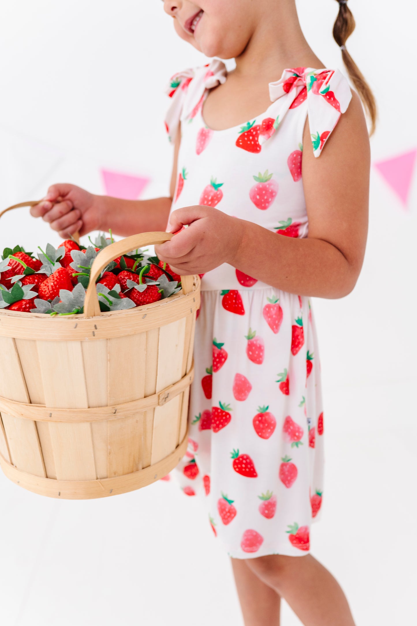 Child in a strawberry print outfit holding a basket of strawberries on a white background