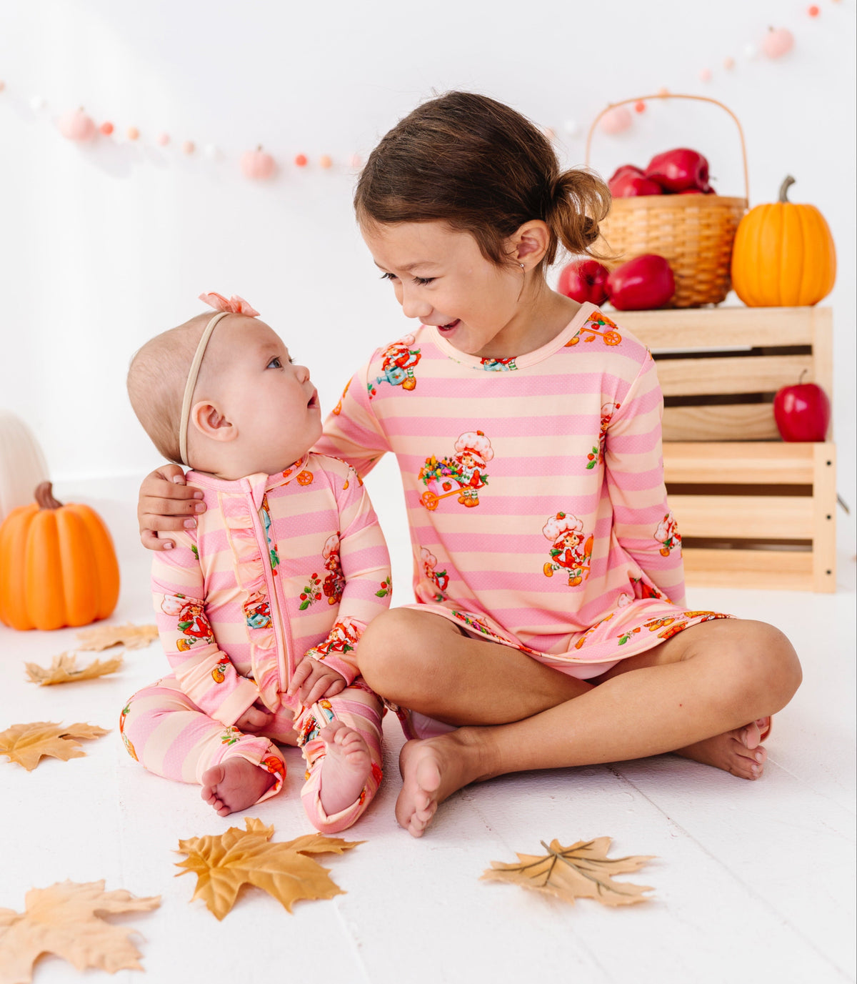 Two children sitting together in a bright indoor setting, wearing matching pink striped long-sleeve pajamas with a fall harvest print of pumpkins, leaves, and apples. Decorative pumpkins, a wooden crate with red apples, and autumn leaves are scattered around them.