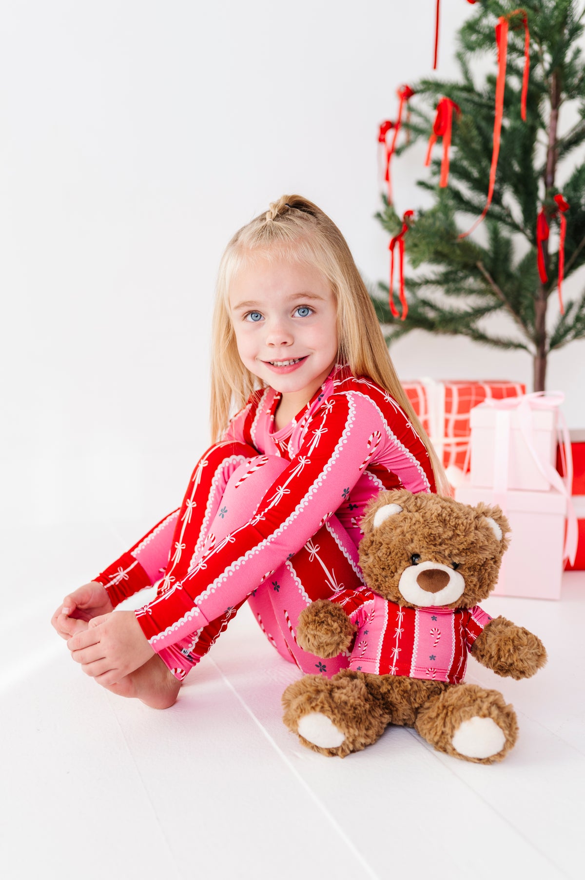 A young girl with blonde hair sits on a white floor next to a teddy bear, wearing a pink and red striped bamboo holiday pajama set. In the background, a Christmas tree with red ornaments and wrapped gifts are visible against a white wall.