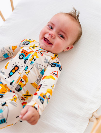 A baby lying on a white muslin mattress in a wooden crib, wearing a cream-colored footed sleeper with a colorful construction vehicle print.