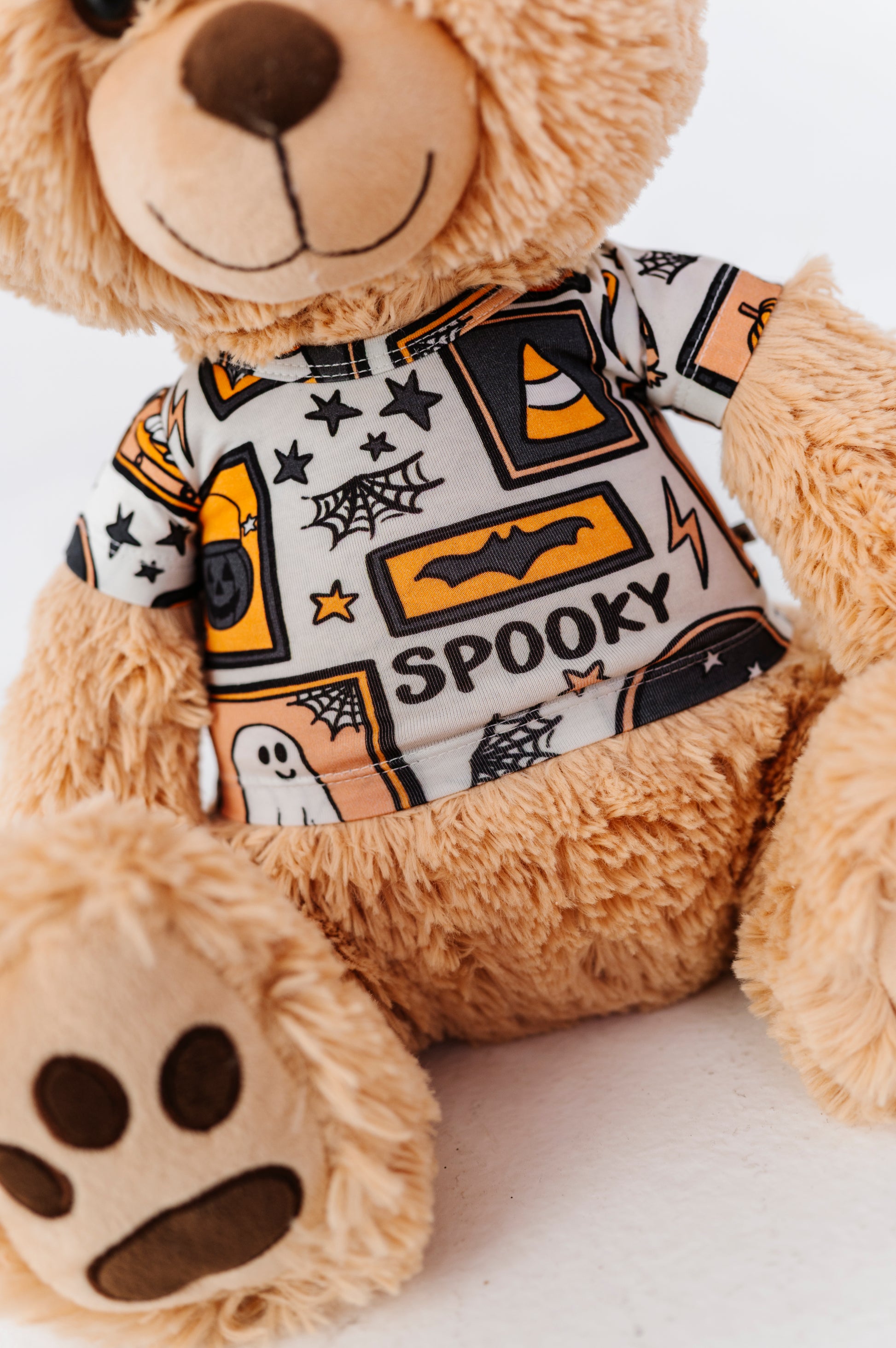 A close-up of a plush teddy bear wearing a short-sleeve shirt with a Halloween-themed print. The shirt features orange, black, and white graphics including the word 'SPOOKY' and spiderwebs, shown against a plain white background.