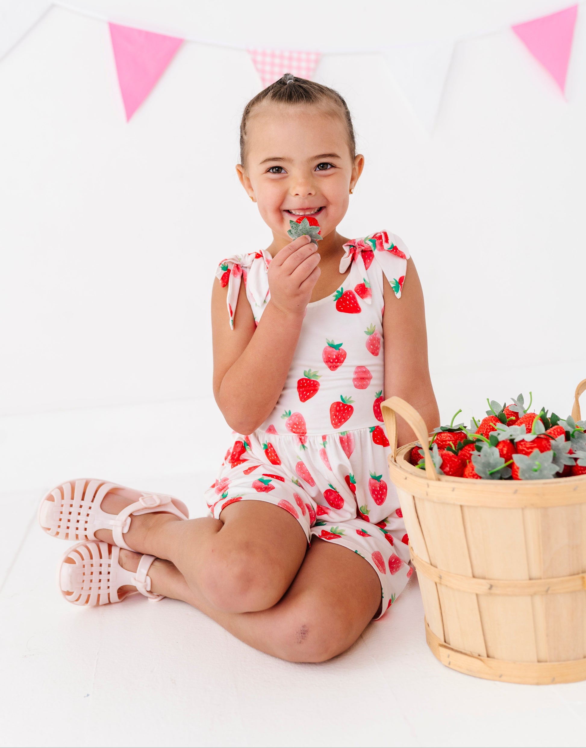 Child in a strawberry-themed outfit sitting next to a basket of strawberries with pink triangle flags in the background.