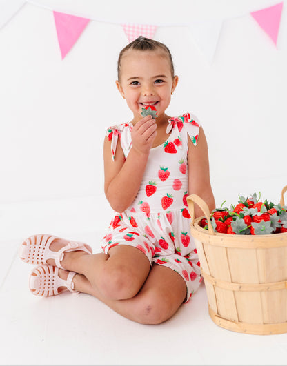 Child in a strawberry-themed outfit sitting next to a basket of strawberries with pink triangle flags in the background.