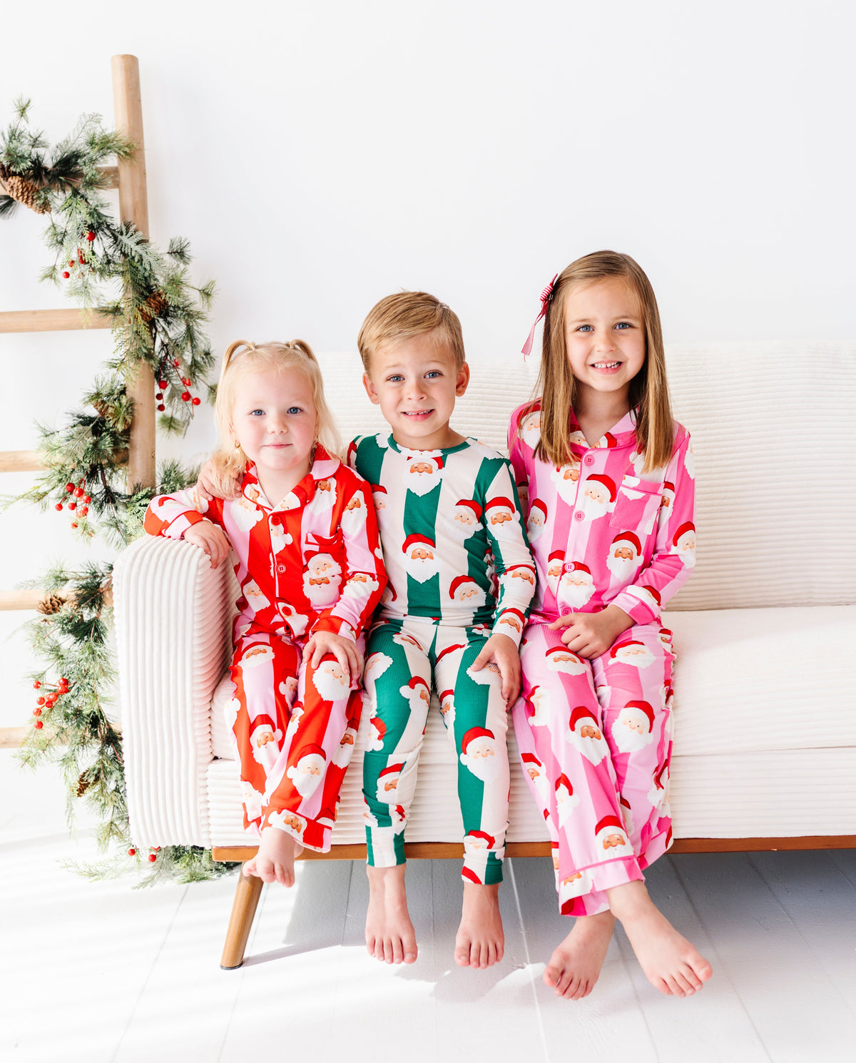 Three children sitting together on a couch in a bright white room with Christmas decor. Their pajamas coordinate with red stripes, green stripes, and pink stripes - all featuring Santa's face 