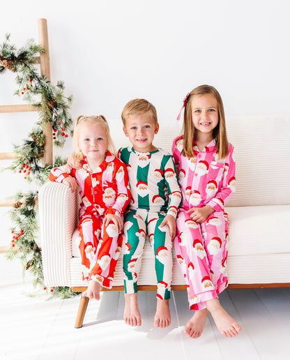 Three children sitting together on a couch in a bright white room with Christmas decor. Their pajamas coordinate with red stripes, green stripes, and pink stripes - all featuring Santa's face 