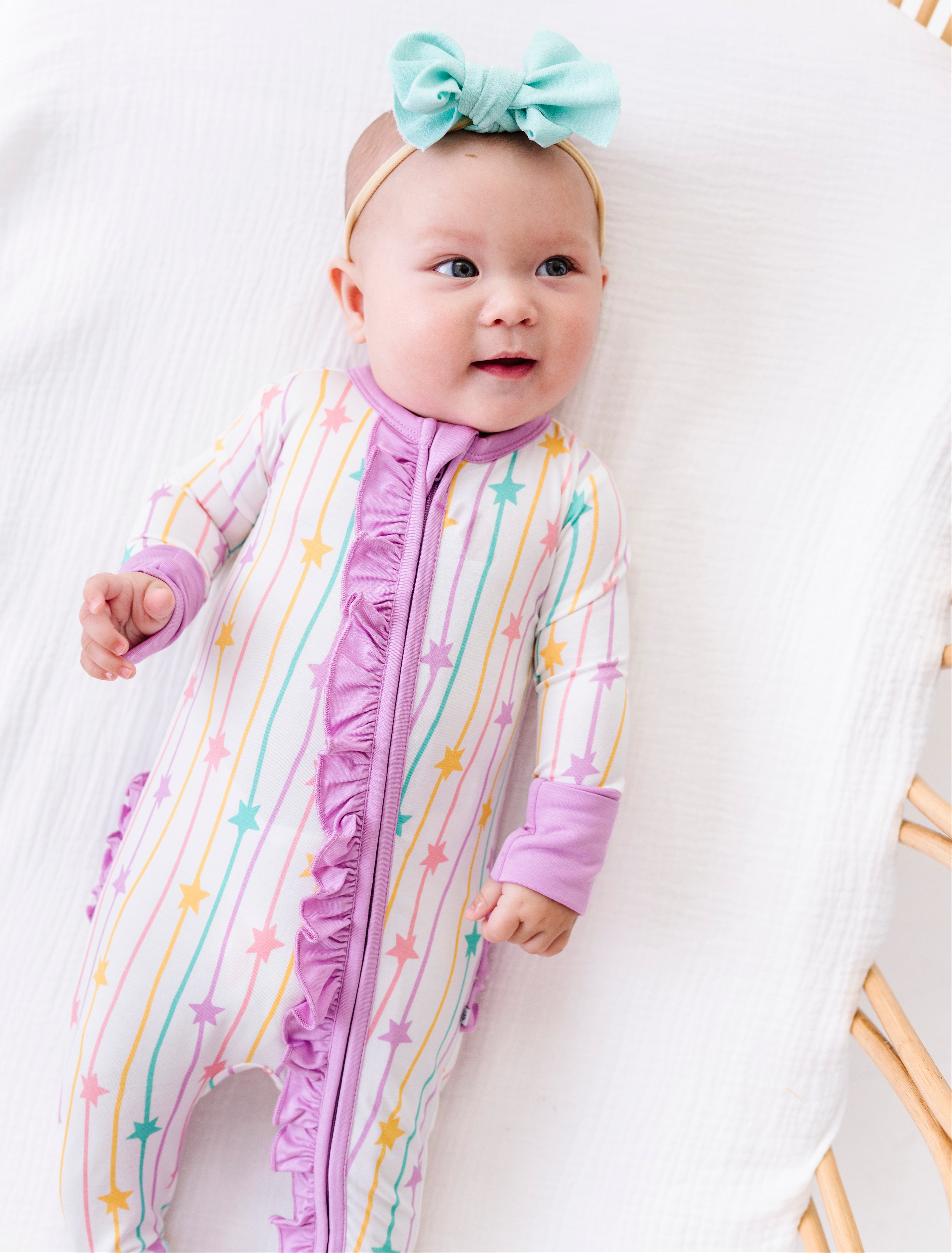 A baby lying on a white muslin bassinet wearing a pastel rainbow striped footless sleeper with purple ruffle trim and a mint green bow headband.