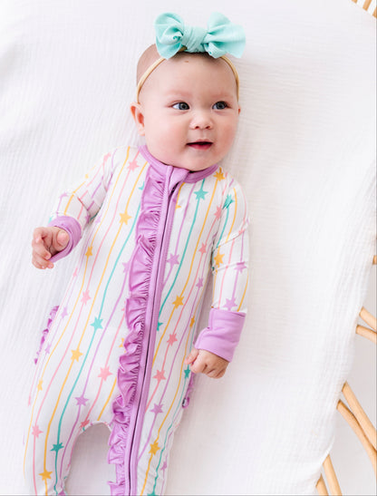 A baby lying on a white muslin bassinet wearing a pastel rainbow striped footless sleeper with purple ruffle trim and a mint green bow headband.