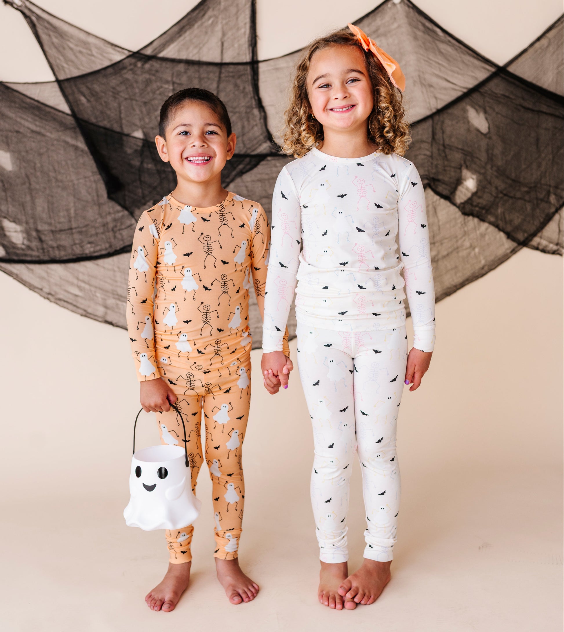 Two children in Halloween-themed pajamas standing in front of a decorative spider web.