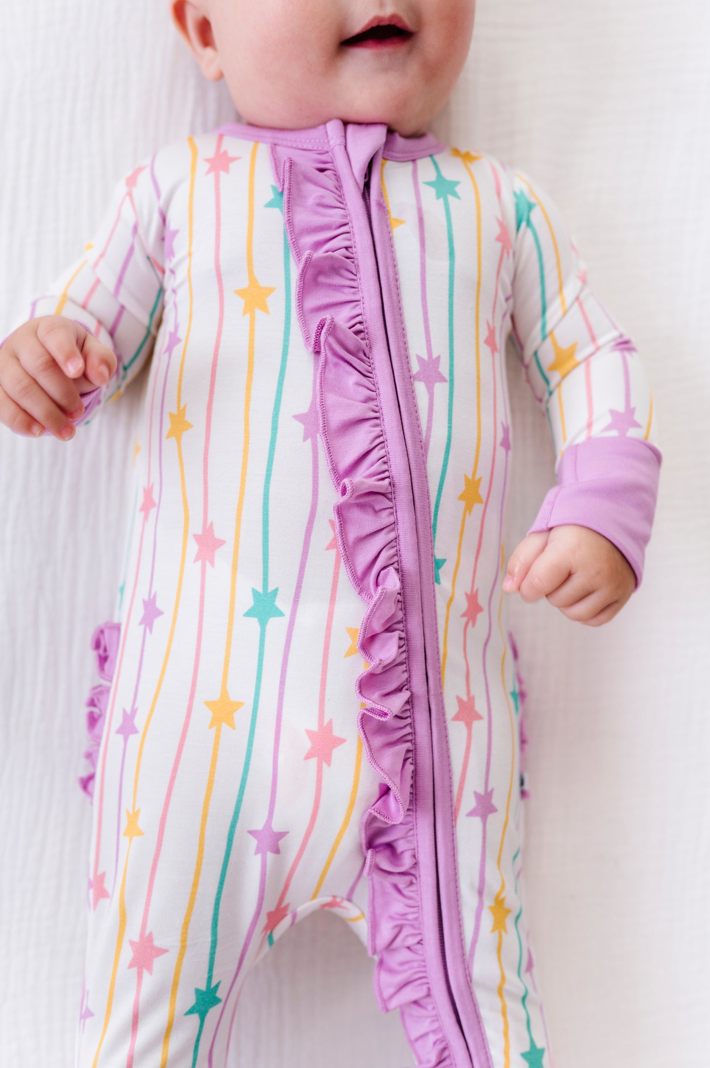 A baby wearing a long-sleeve footless sleep sack with a vertical ruffle zipper. The sleep sack has a white base with vertical pastel stripes in pink, yellow, and teal, accented with pastel pink and teal stars.