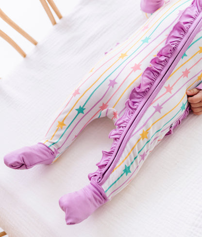 A baby's legs wearing a footed sleeper with a pastel rainbow star and stripe pattern, featuring a purple ruffle zipper and matching purple foot cuffs, resting on a white textured blanket.
