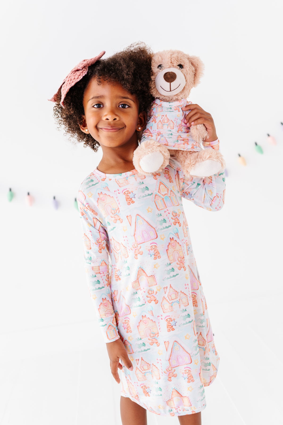 A little girl wearing a Christmas gingerbread-village themed bamboo gown and holding a 13" teddy bear wearing a matching bamboo gown in the same Christmas print 