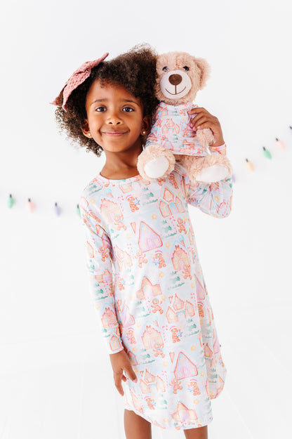 A little girl wearing a Christmas gingerbread-village themed bamboo gown and holding a 13" teddy bear wearing a matching bamboo gown in the same Christmas print 
