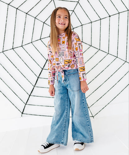 A young girl stands in front of a white studio backdrop with a large black spiderweb graphic, wearing a pink long-sleeve top with a colorful food-themed patchwork print, wide-leg light blue jeans, and black-and-white sneakers.