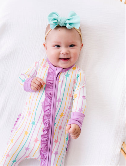 A smiling baby lying on a white muslin mattress in a wooden crib, wearing a pastel rainbow star-patterned footless sleeper with purple ruffle trim and a mint green bow headband.