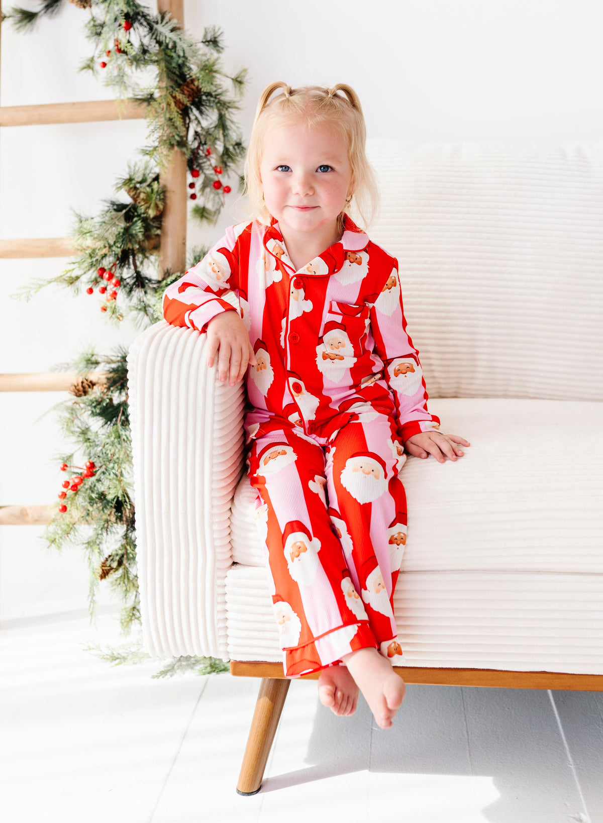 A young girl with blonde pigtails sits on a white armchair wearing a red and pink Santa Claus bamboo pajama set. The background features a white wall, a wooden ladder, and a green holiday garland with red berries.