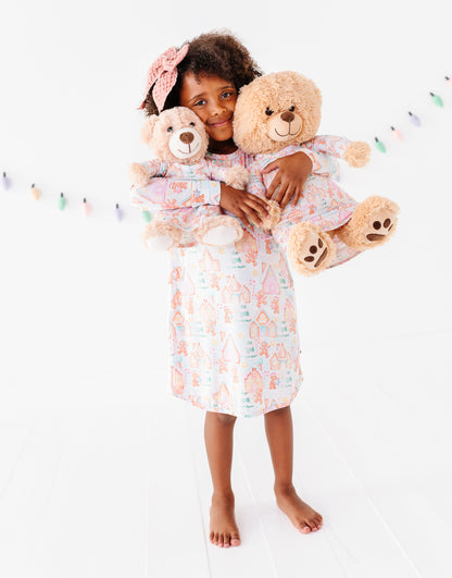 A little girl standing in a bright white room wearing a bamboo Christmas-themed gingerbread village gown and holding two stuffed animals wearing matching bamboo gowns 
