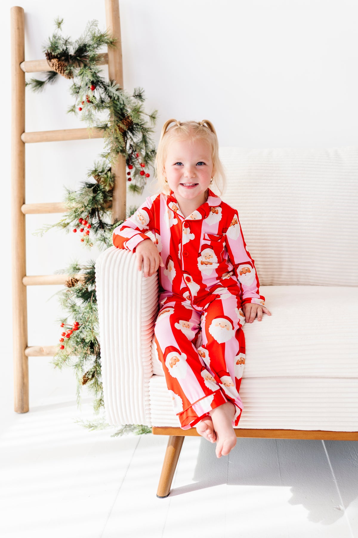 A young girl with blonde pigtails sits on a white armchair wearing a red and white Santa Claus patterned bamboo pajama set. The scene is decorated with a wooden ladder and green holiday garland with red berries against a bright white background.