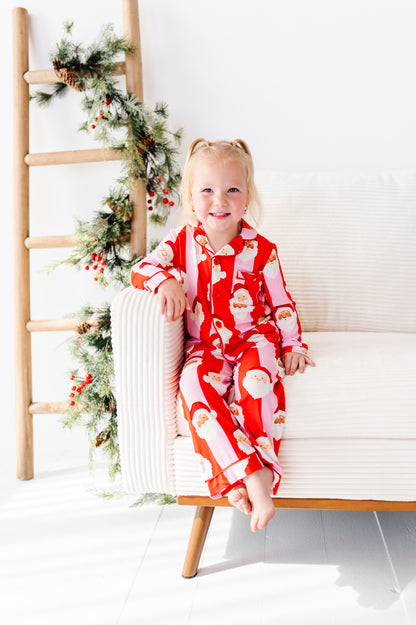 A young girl with blonde pigtails sits on a white armchair wearing a red and white Santa Claus patterned bamboo pajama set. The scene is decorated with a wooden ladder and green holiday garland with red berries against a bright white background.