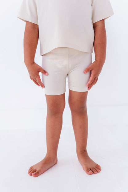 Little girl wearing ivory biker shorts on a plain background