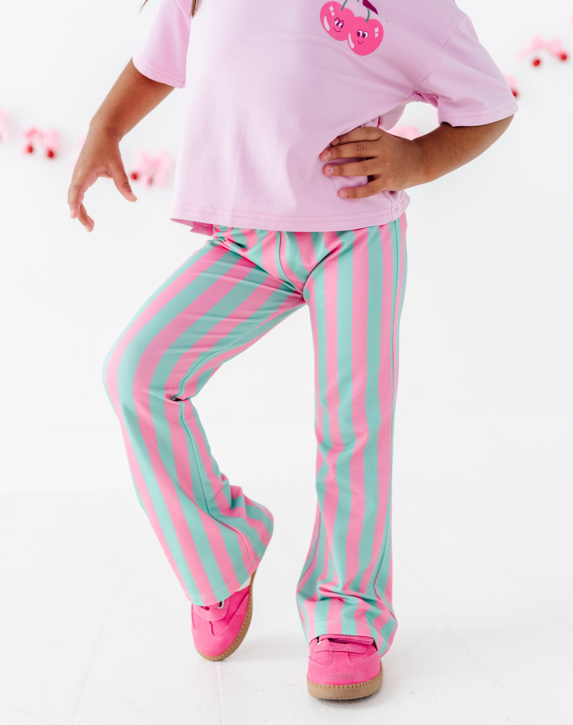 A child wearing pink and mint green vertically striped pants, a light pink t-shirt with a cherry graphic, and pink shoes, shown from the waist down against a white studio background.