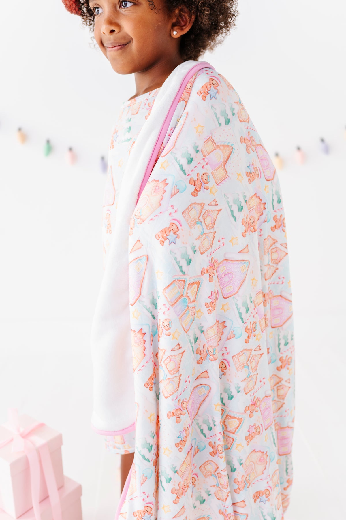 A little girl wearing a bamboo nightgown holding a matching Christmas plush blanket featuring a gingerbread village 