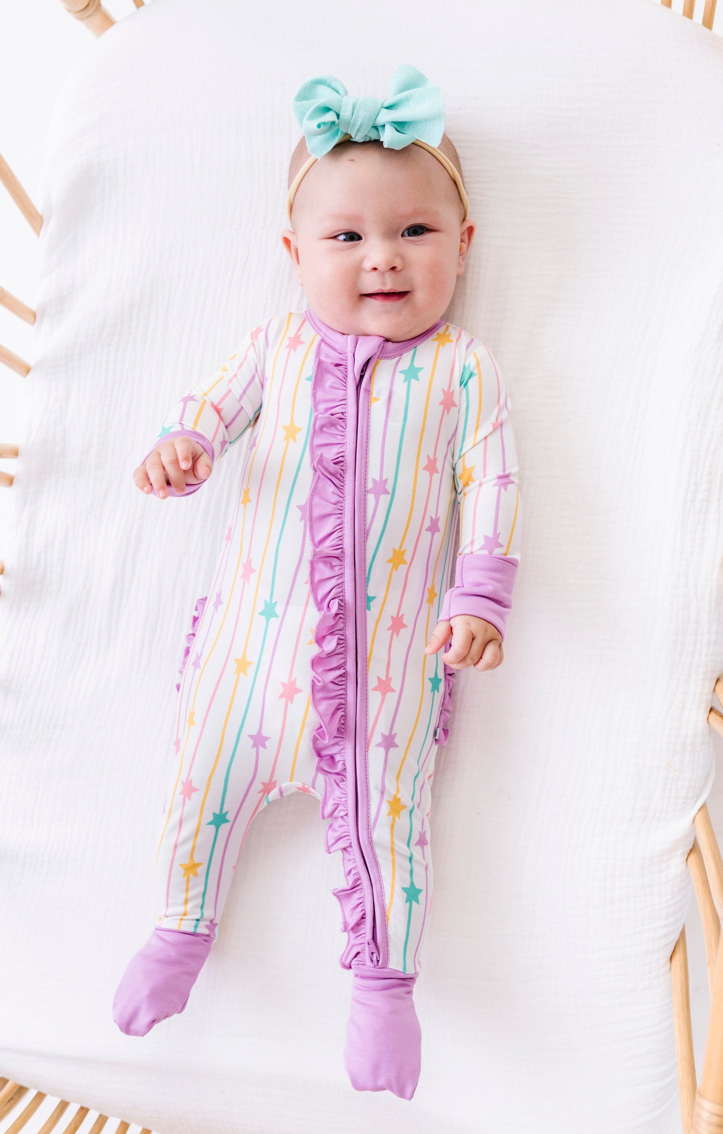 A baby lying on a white muslin mattress inside a wicker bassinet, wearing a pastel rainbow striped footed sleeper with purple ruffle trim and a mint green bow headband.