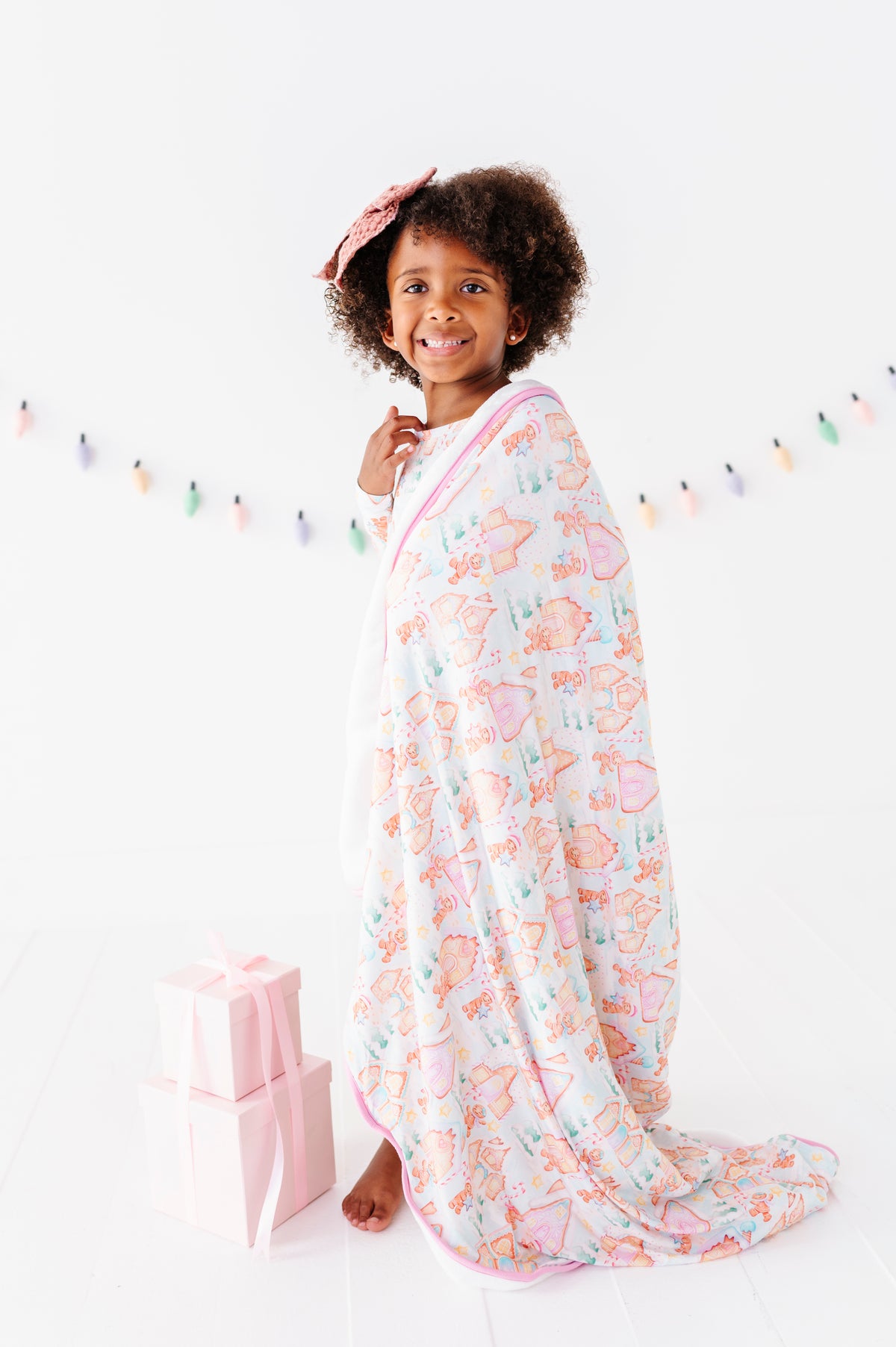 A little girl wearing a Christmas gingerbread village-themed gown, standing in a bright white room and wrapped in a plush bamboo gown in a matching gingerbread village print 