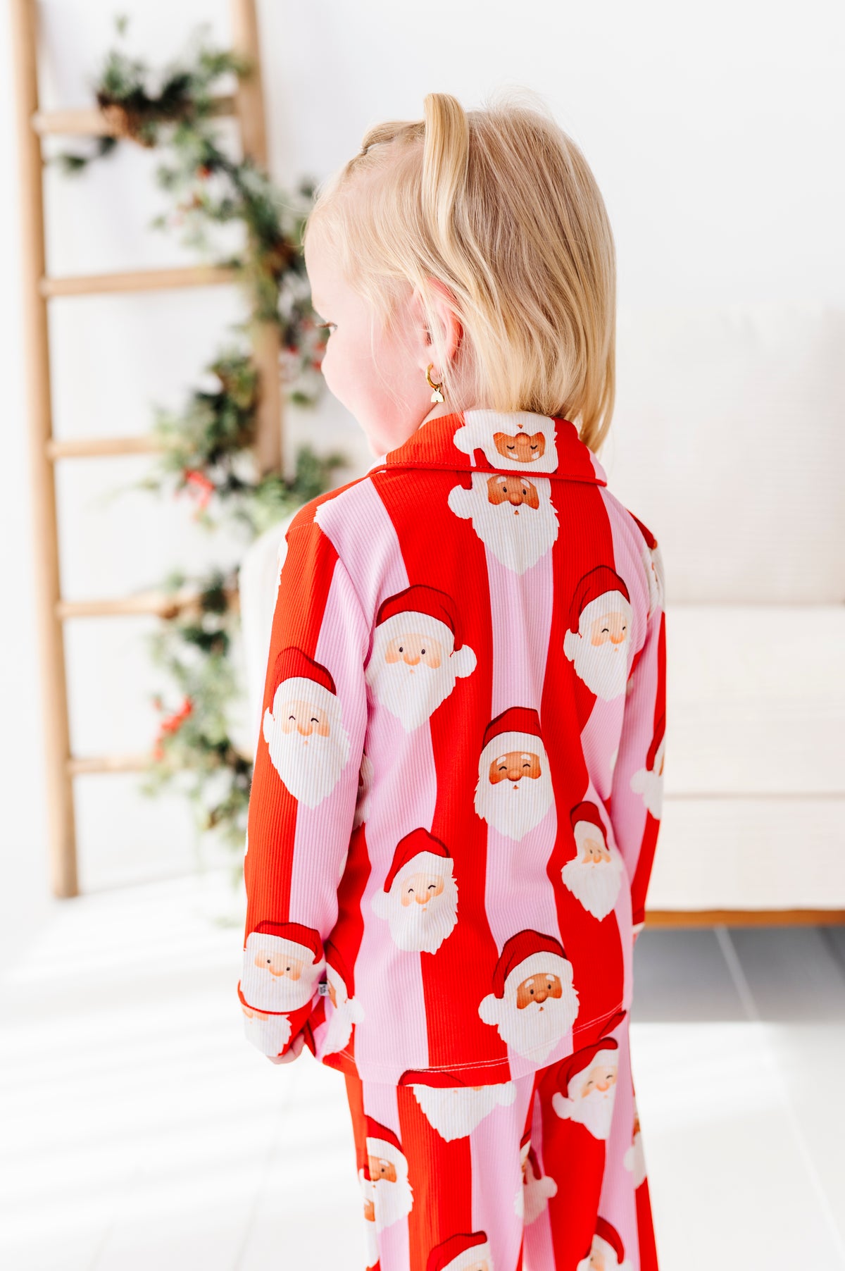 A young child viewed from behind wearing a red and pink Santa Claus patterned bamboo pajama set in a bright indoor room with a ladder and holiday decor in the background.