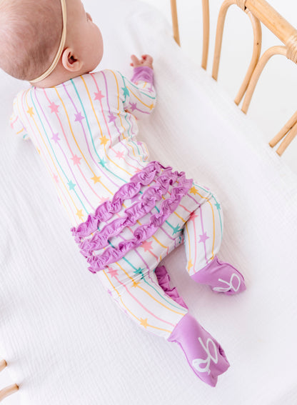 A baby lying on their side on a white textured blanket inside a wooden crib, wearing a white footed sleeper with pastel rainbow stripes, a purple ruffled waistband, and purple socks.