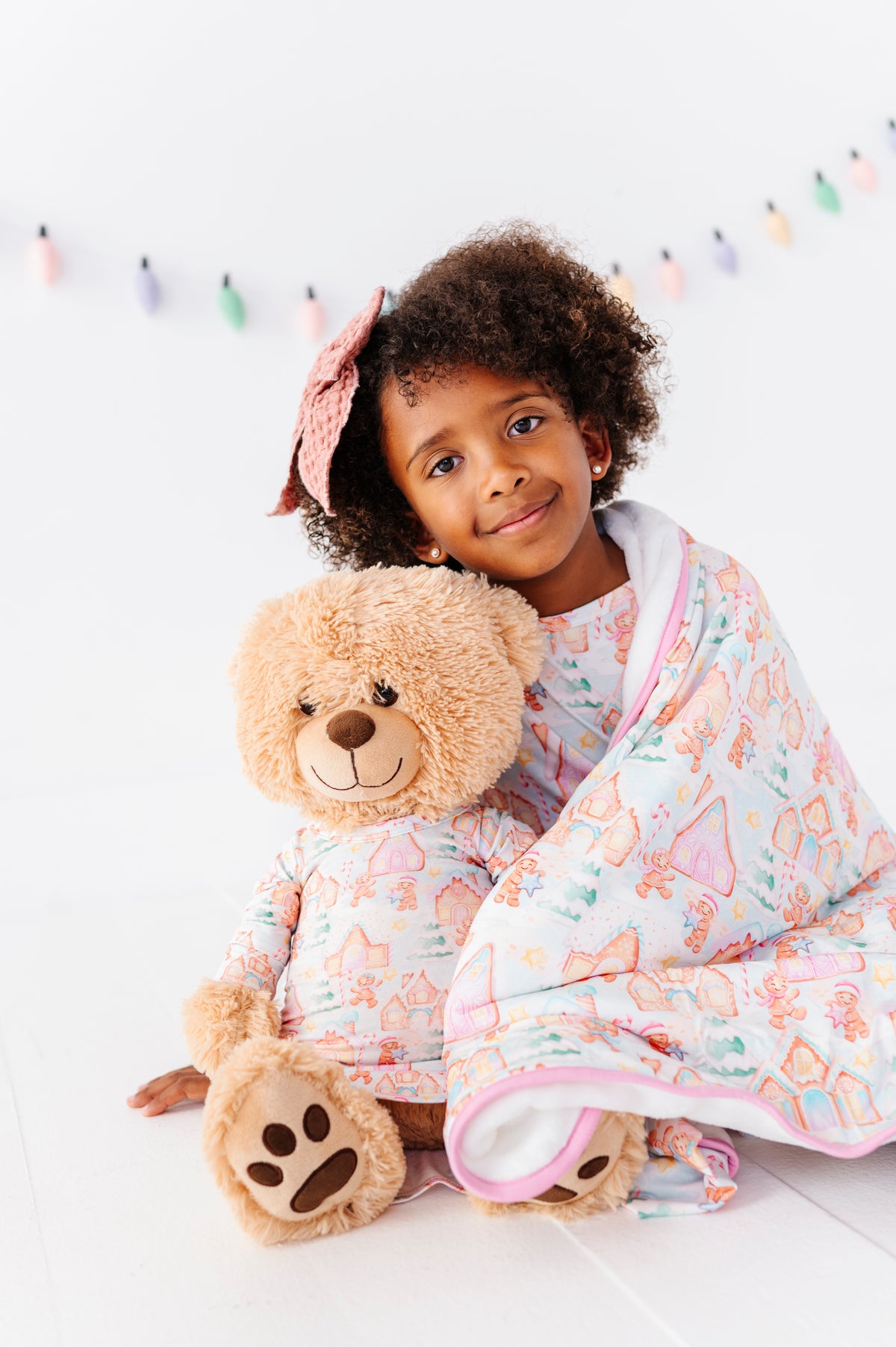 A little girl wearing a bamboo gown featuring a Christmas gingerbread village-themed print and cuddling with her stuffed teddy bear in the same print 