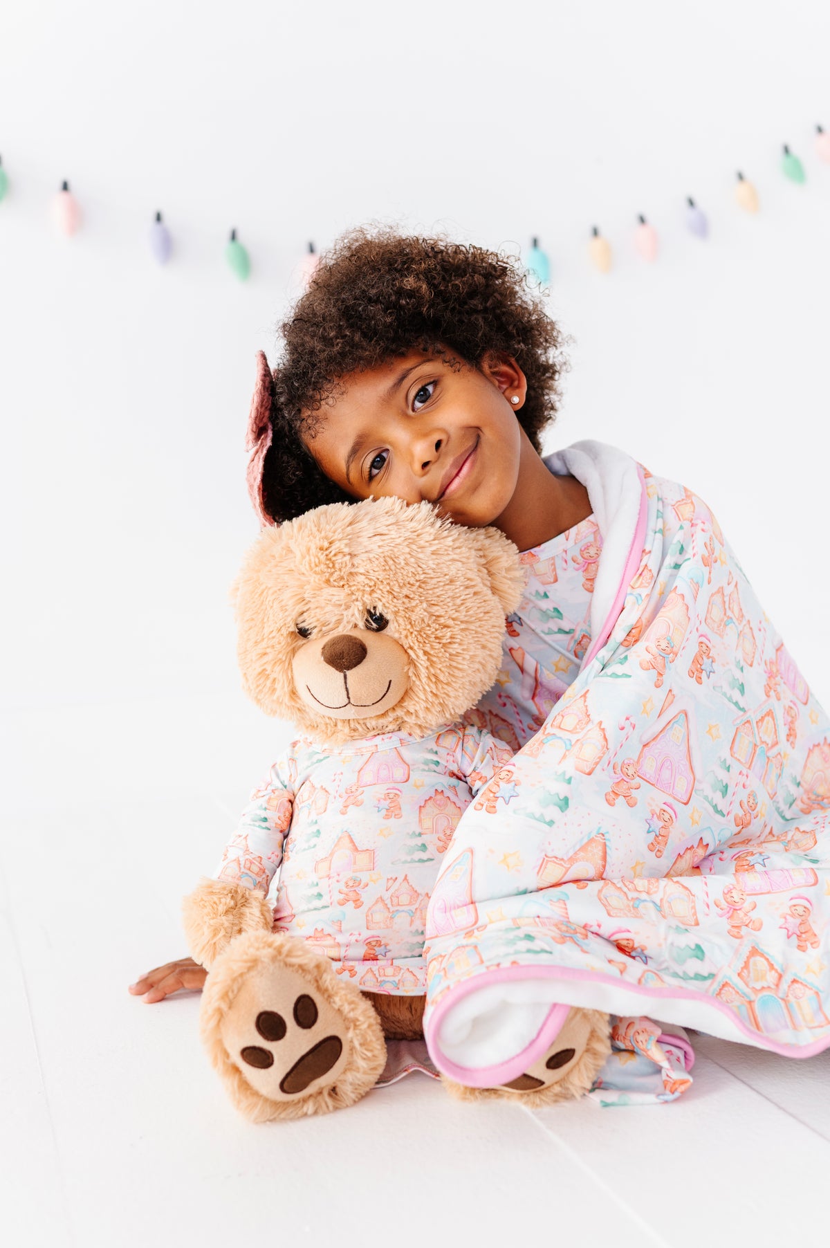 A little girl sitting on the floor and cuddling with her teddy bear. Both the little girl and the stuffy are wearing matching Christmas bamboo gowns, and they're wrapped in a Christmas gingerbread village bamboo plush blanket 
