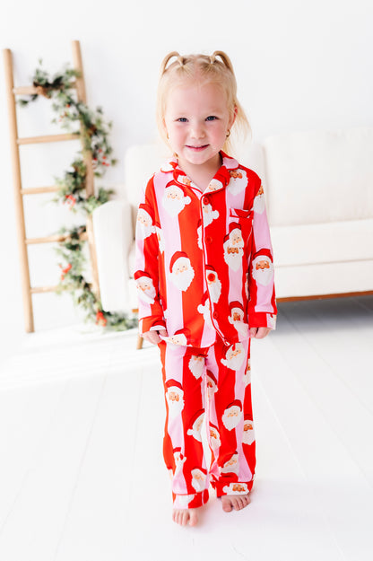 A young girl with blonde pigtails stands barefoot in a bright room wearing a red and white Santa Claus patterned bamboo pajama set.
