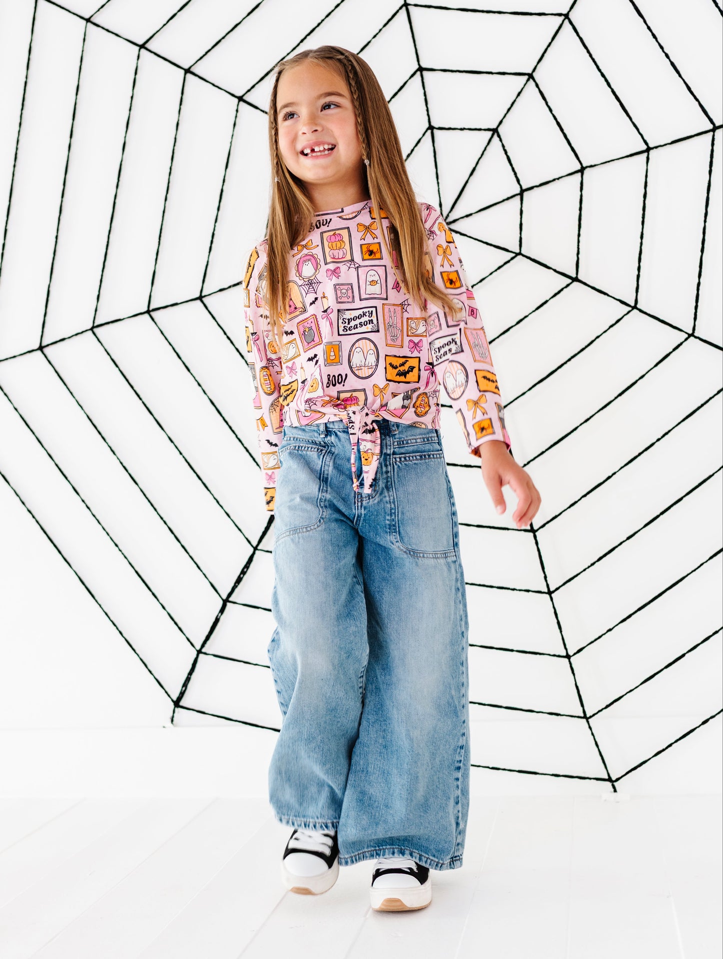 A young girl stands in a studio wearing a pink long-sleeve top with a colorful patchwork print and wide-leg light blue jeans, posed in front of a large black spiderweb graphic on a white background.