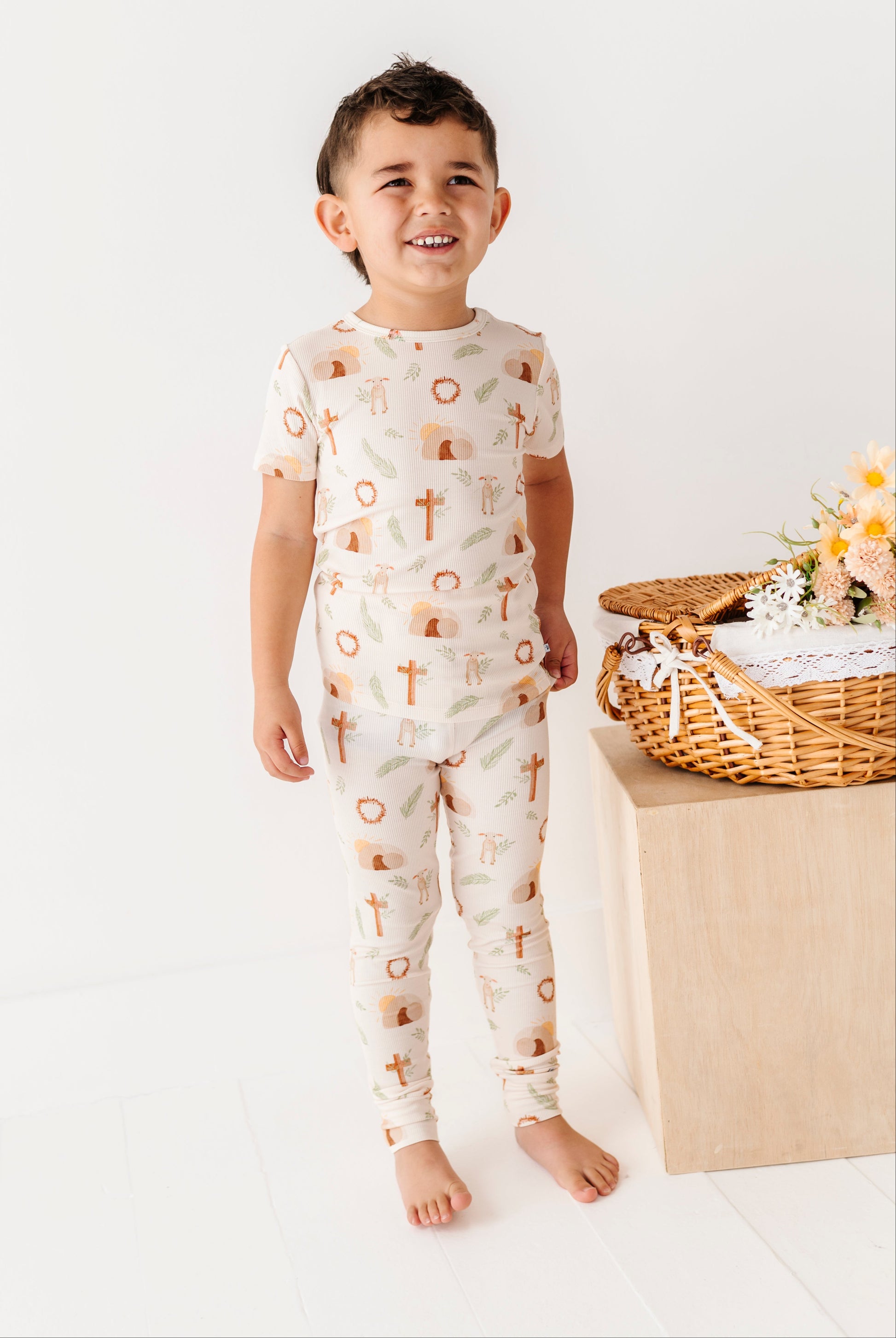 Child wearing a patterned onesie standing in a minimalistic setting with a basket and flowers.
