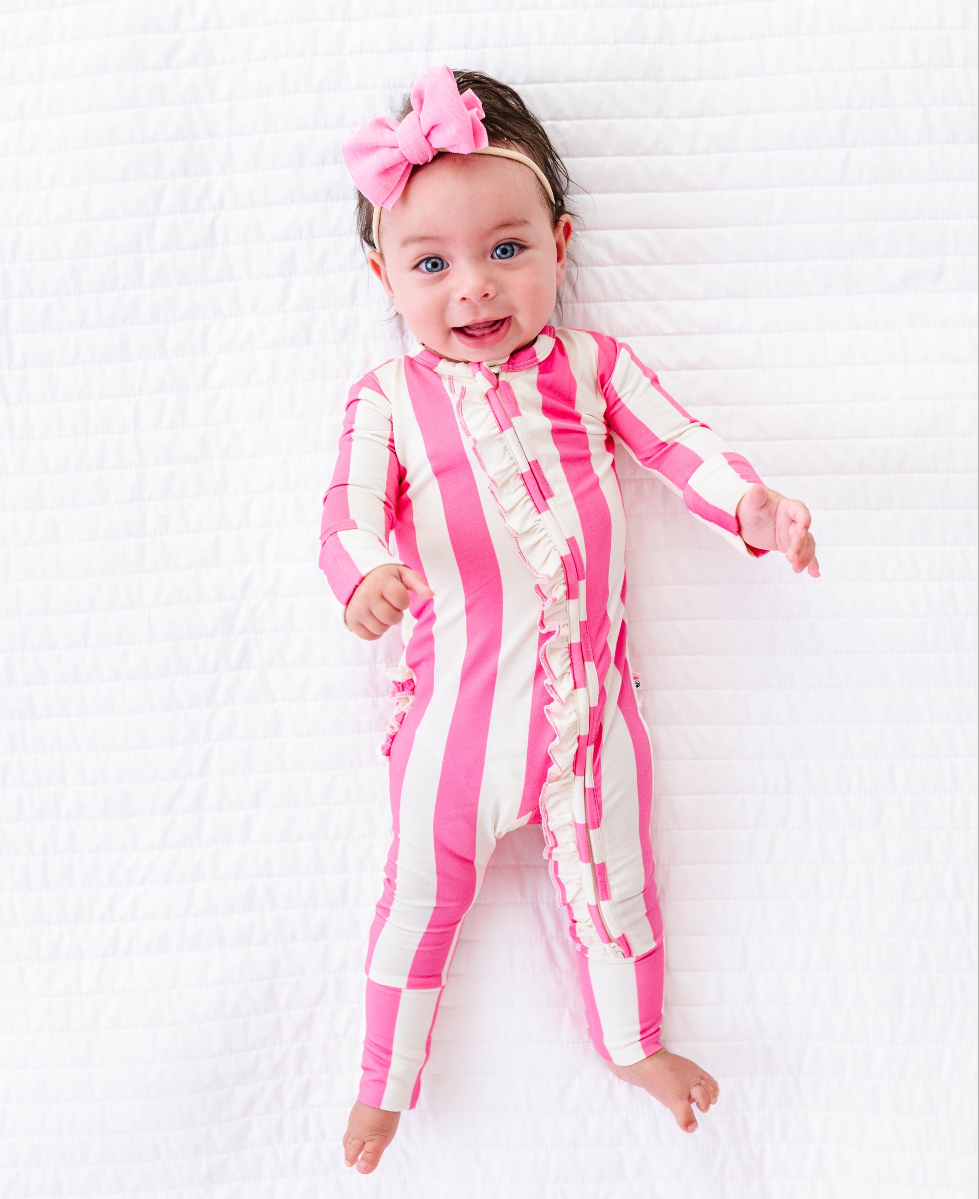 A baby lying on a white textured blanket, wearing a pink and white vertically striped ruffle jumpsuit and a matching pink bow headband.
