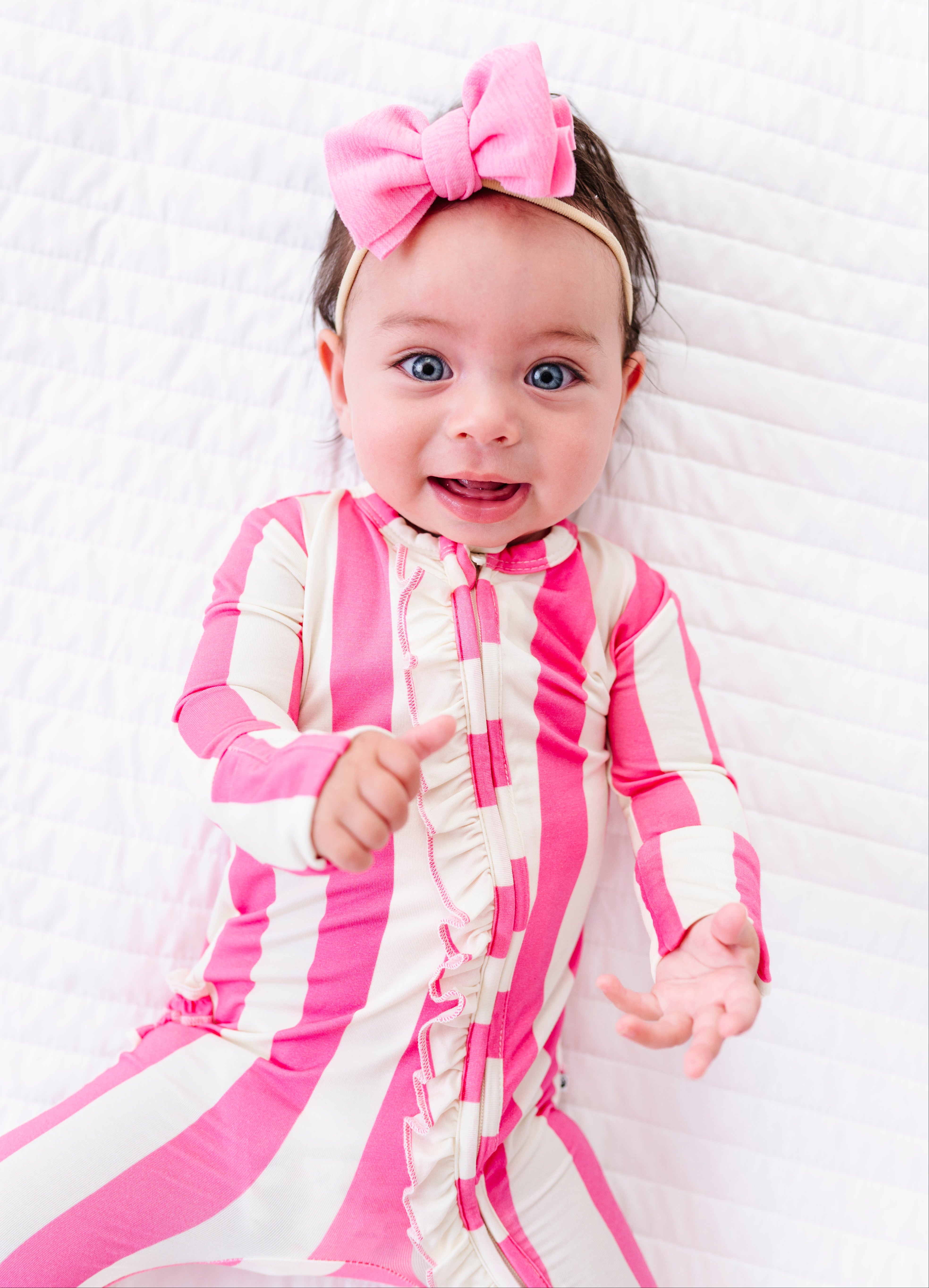 A baby girl wearing a pink and white striped long-sleeve bodysuit and a matching pink bow headband, lying on a white quilted blanket.