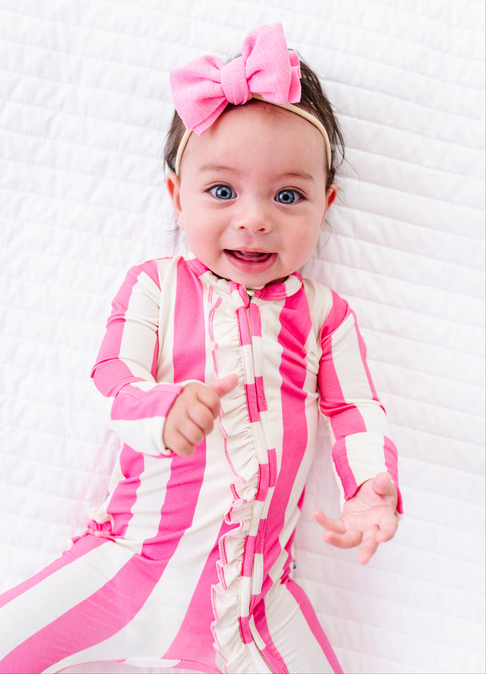 A baby girl wearing a pink and white striped long-sleeve bodysuit and a matching pink bow headband, lying on a white quilted blanket.